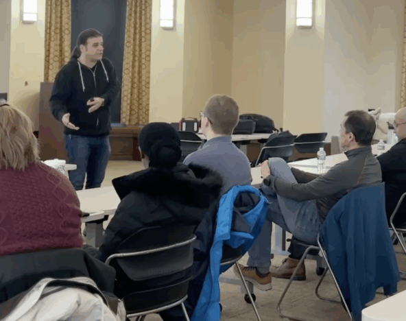A person stands and speaks to a seated group of adults in a classroom or meeting room setting, with tables, chairs, and jackets visible.